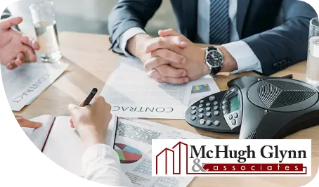 Two professionals shaking hands over a desk with documents, pens, and a conference phone and "McHugh Glynn & associates" logo