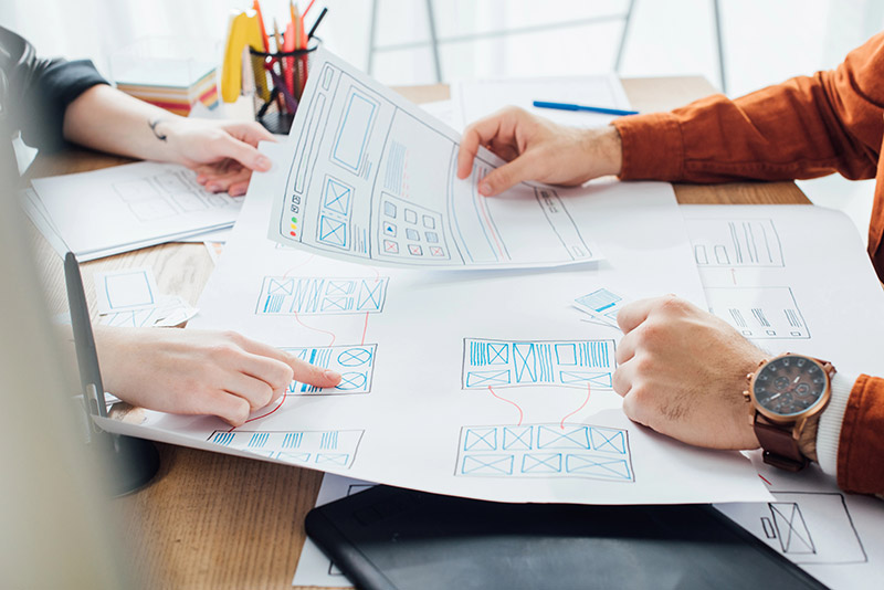 Two people collaborate on UX design, reviewing and pointing at hand-drawn webpage wireframes on a cluttered desk.
